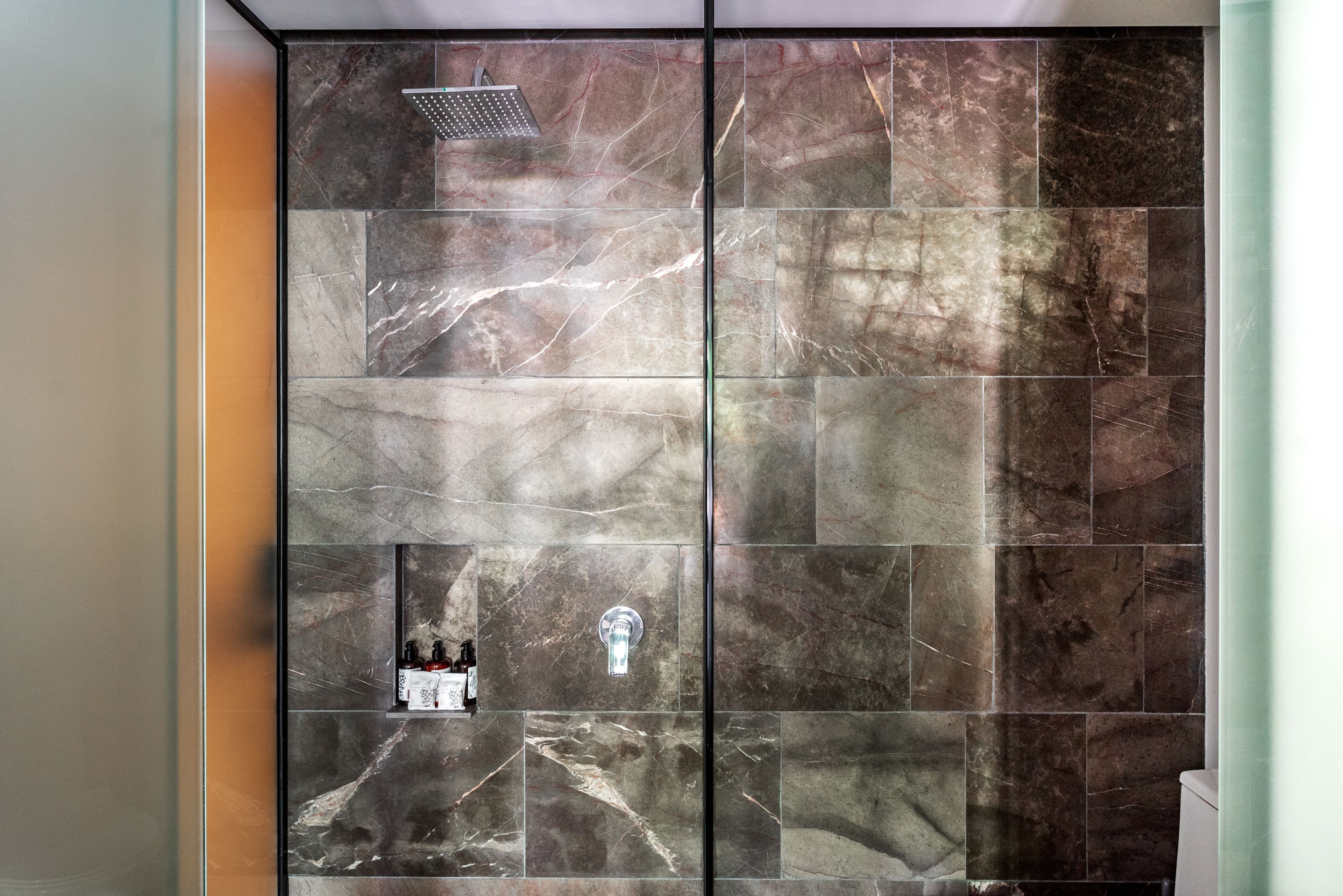 Un baño con zona de ducha, pared de azulejos de mármol y mampara de vidrio.