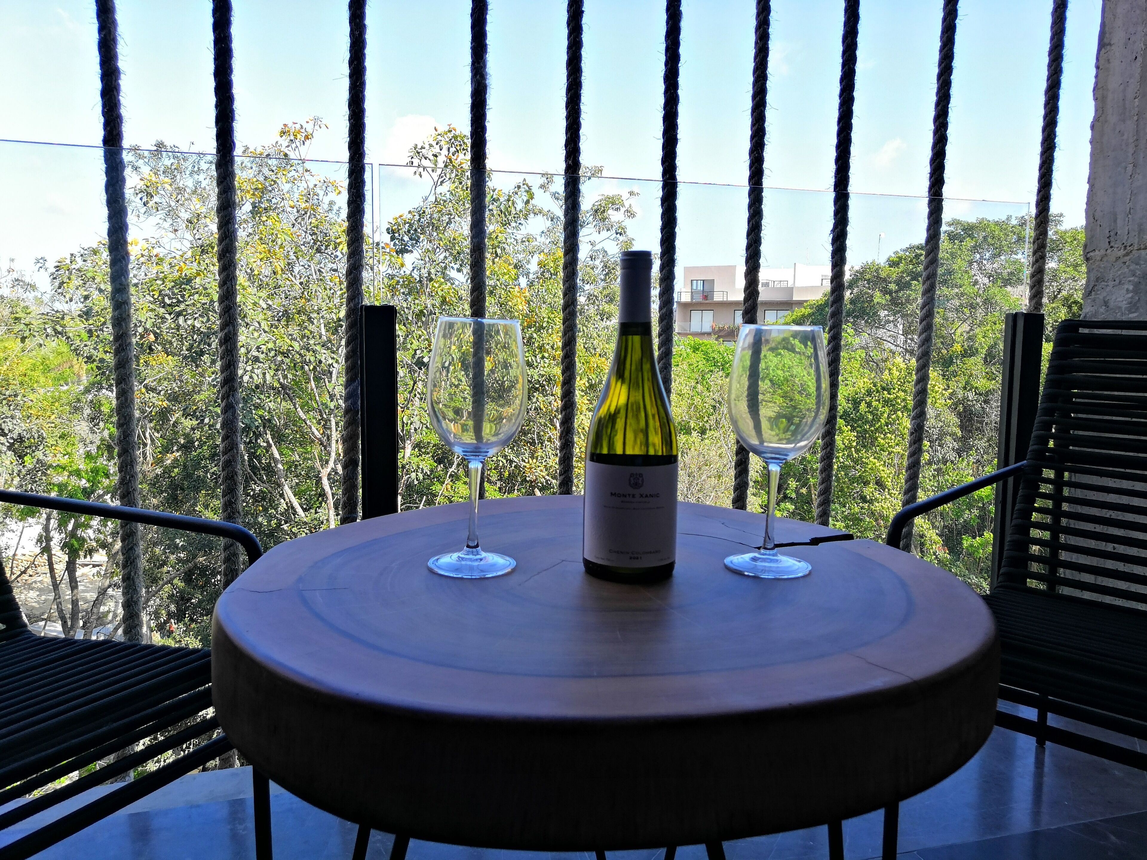 Mesa redonda con una botella de vino y dos copas en un balcón con vistas a árboles y edificios.