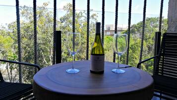 Mesa redonda con una botella de vino y dos copas en un balcón con vistas a árboles y edificios.