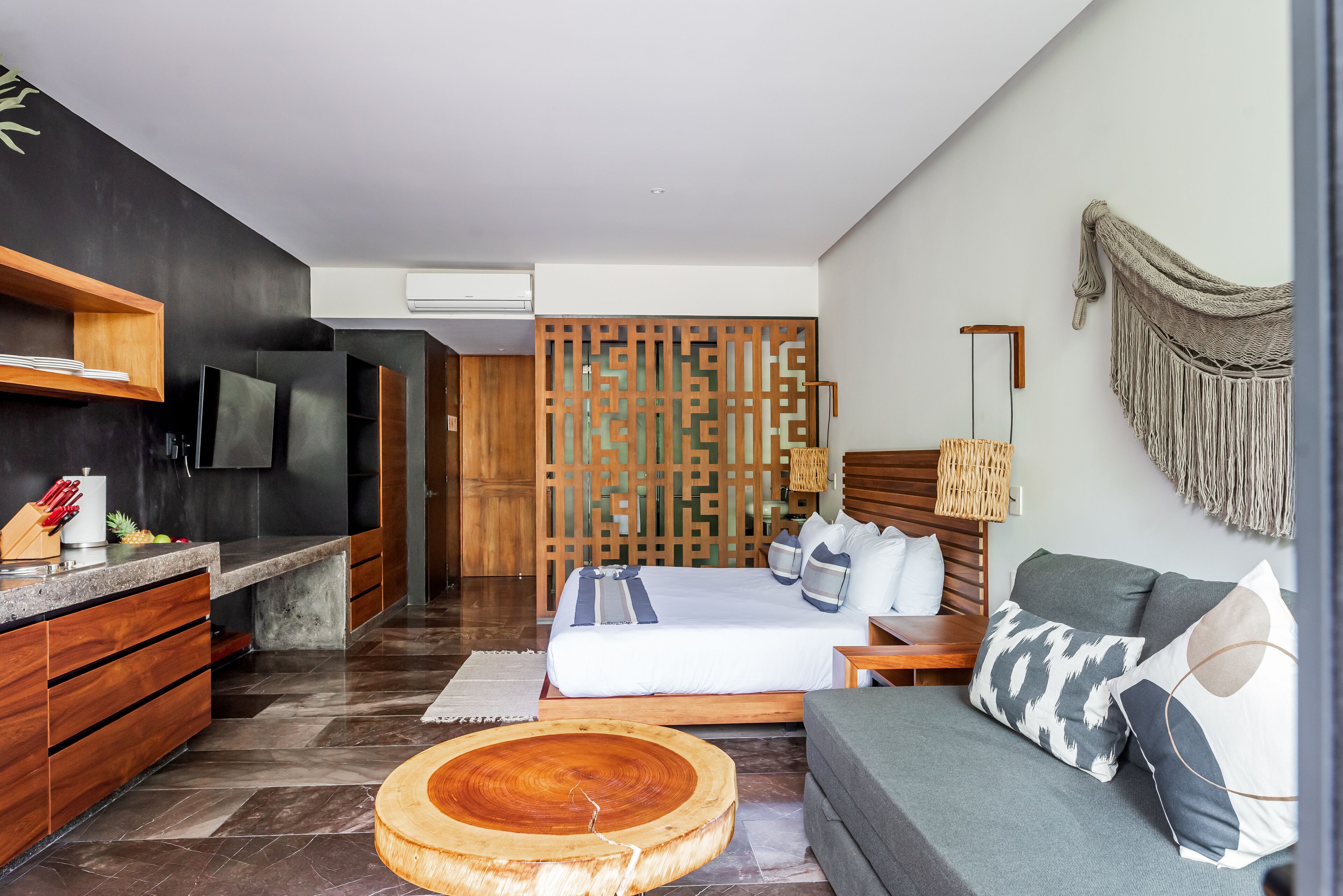 A modern hotel room with a wooden bed, grey sofa, and a kitchen area with a wooden counter and cabinets.