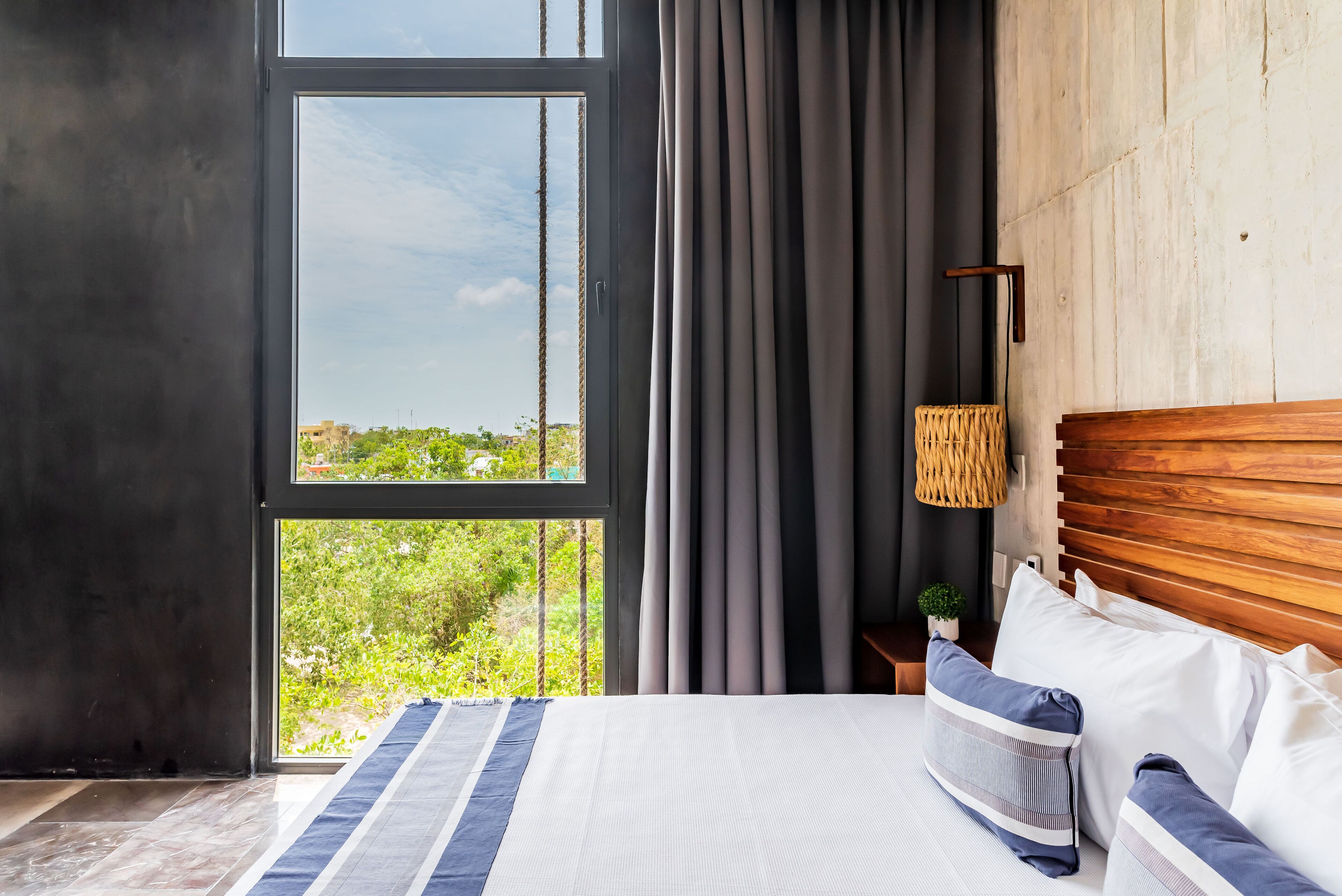 Ein modernes Schlafzimmer mit großem Fenster, einem Bett mit gestreiften Kissen, einem hölzernen Kopfteil und einer kleinen Pflanze auf dem Nachttisch.