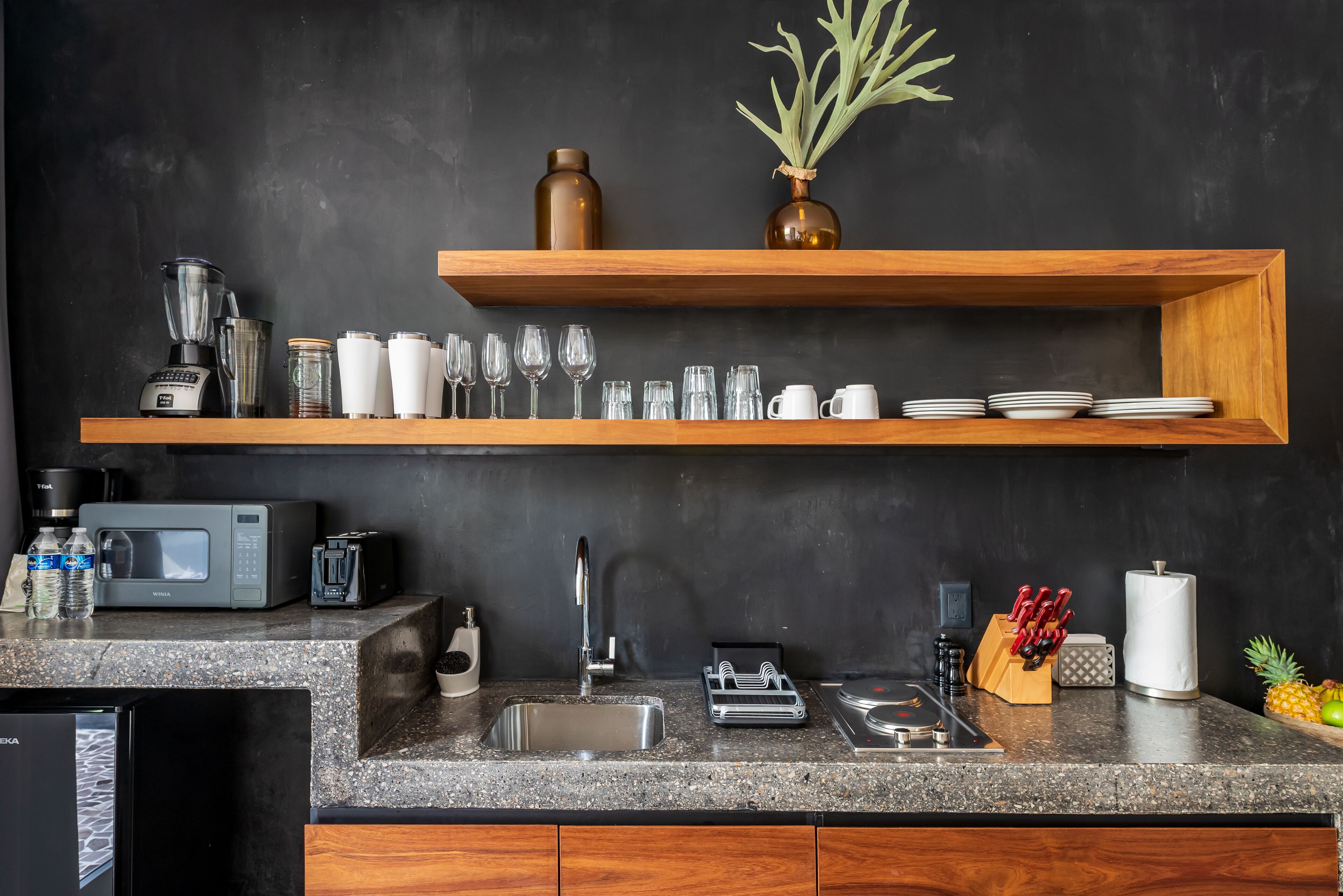 A modern kitchen with granite countertops, a built-in dishwasher, a microwave, and a sink. There are wooden shelves above the counter holding glassware and a vase with greenery.