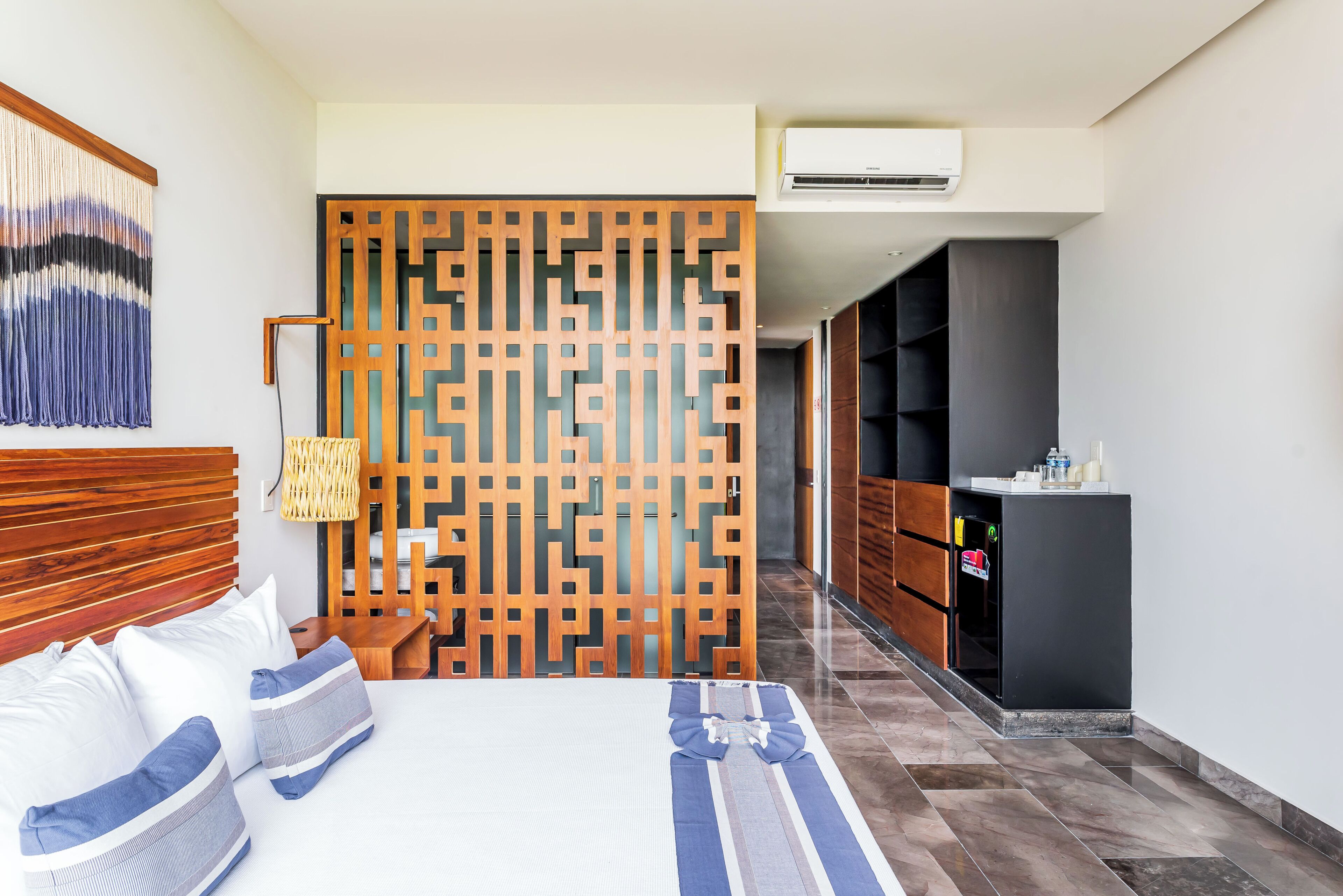 A hotel room with a bed, a wooden partition, a hanging light fixture, and a minibar.