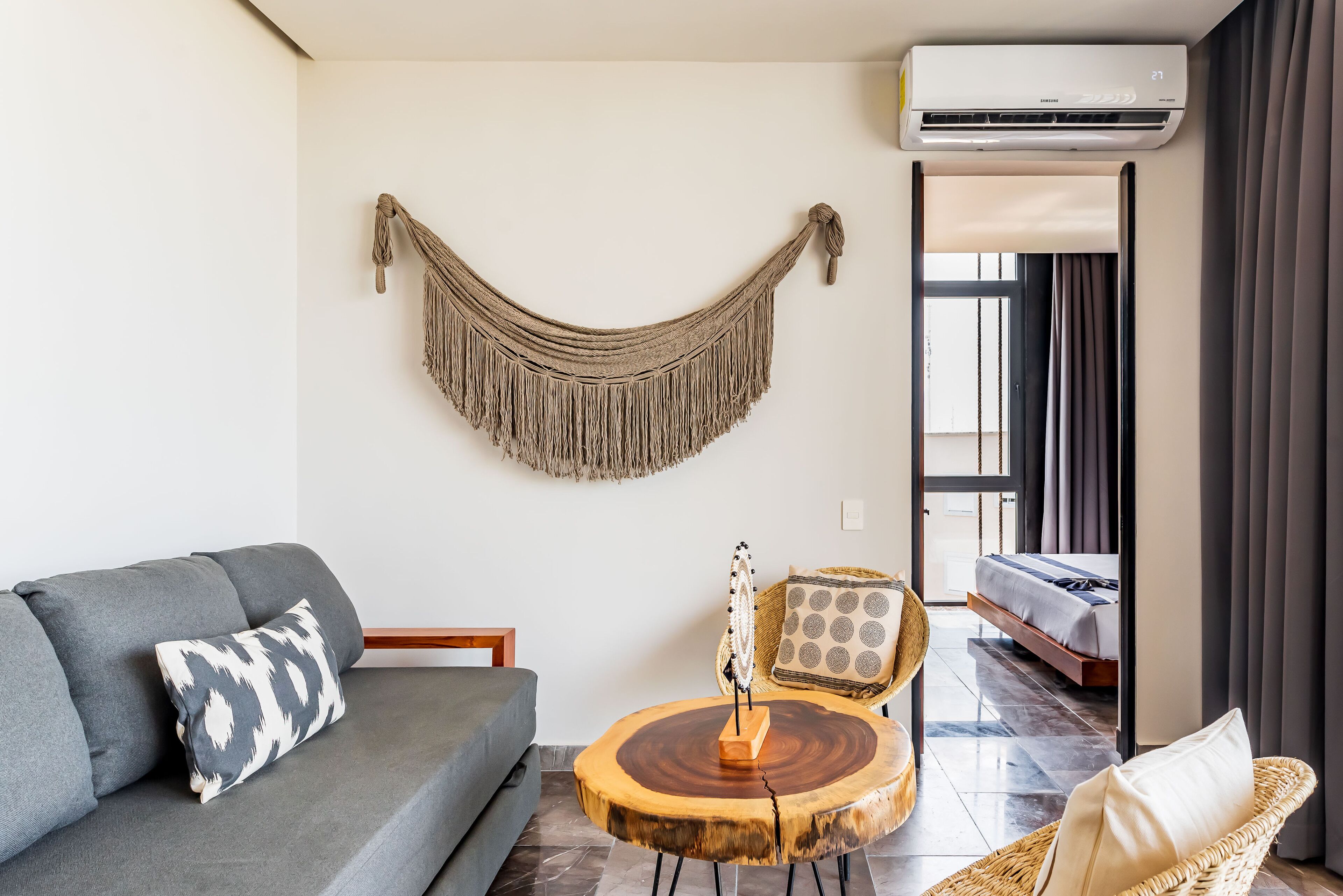 A living room with a gray sofa, a round wooden coffee table, a chair with a woven backrest, and a macramé wall hanging.