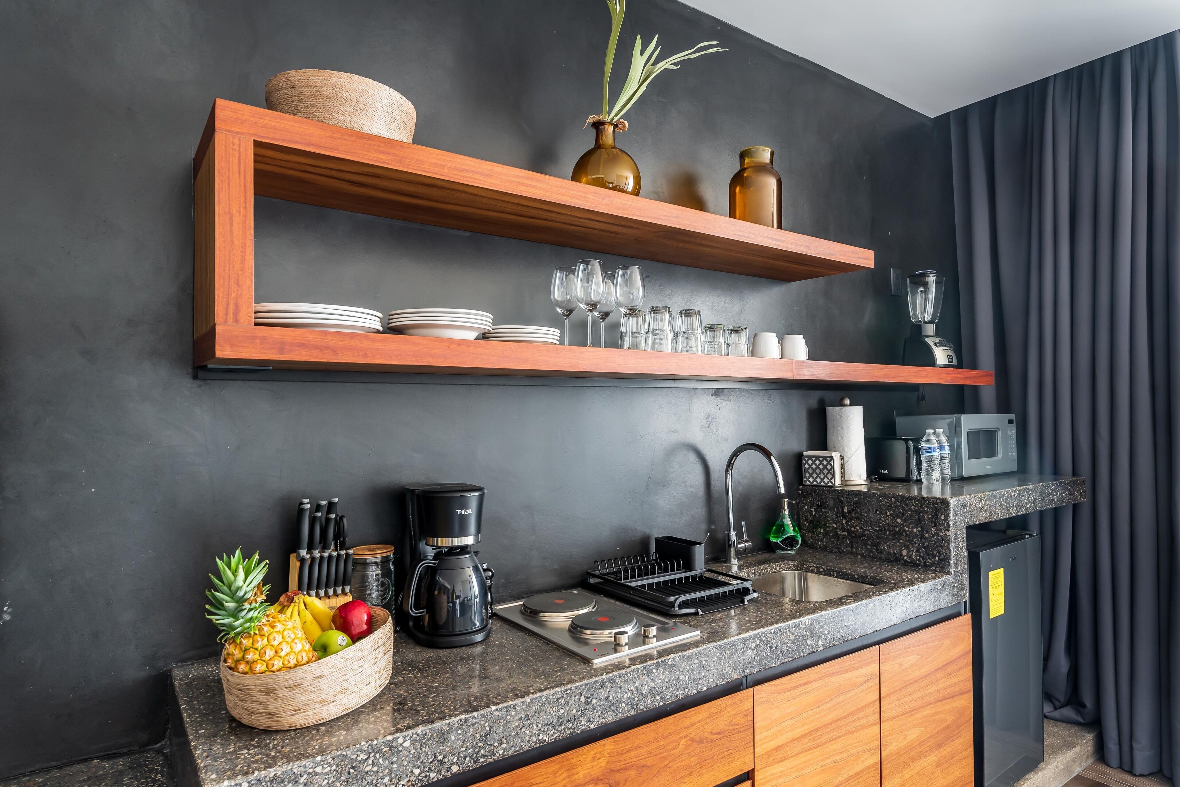 A modern kitchen with wooden shelves, a granite countertop, a coffee maker, and a fruit bowl.