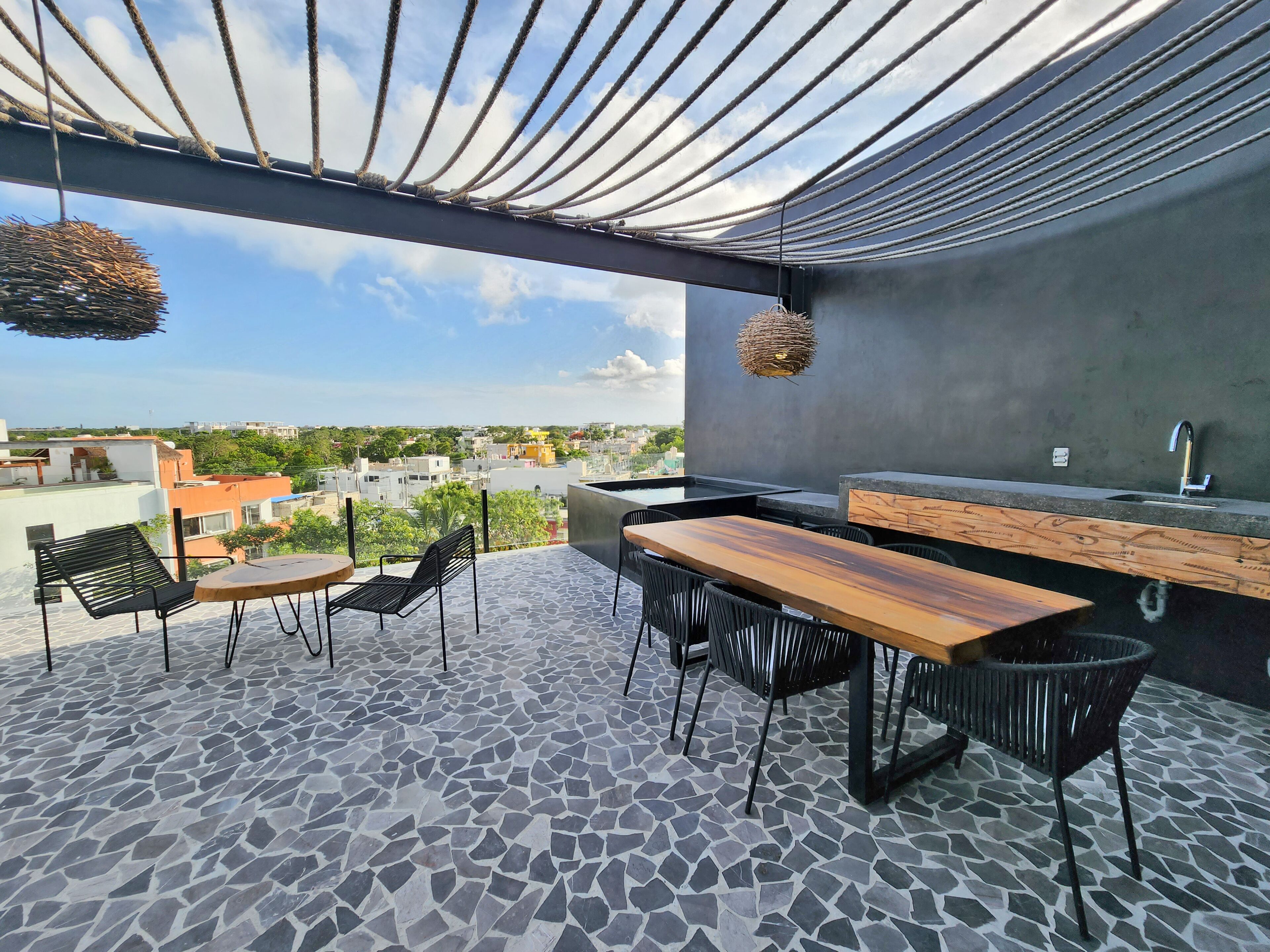 Eine Dachterrasse mit Steinplattenboden, einem Holztisch, schwarzen Stühlen, einer hängenden Leuchte und Blick auf die Stadtlandschaft.