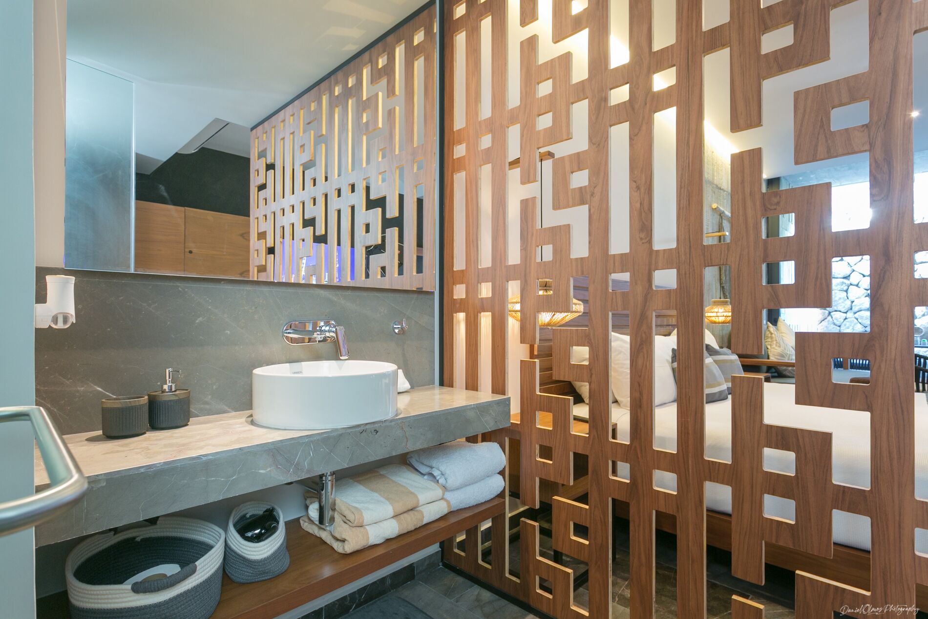 A modern bathroom with a large mirror, a white sink, and a wooden partition with cut-out patterns.