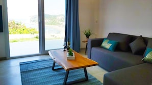 A modern living room with a sofa, a wooden coffee table, and a view of a coastal landscape through a large window. - Gikas Apartments - Serenity Suite (Corfu)