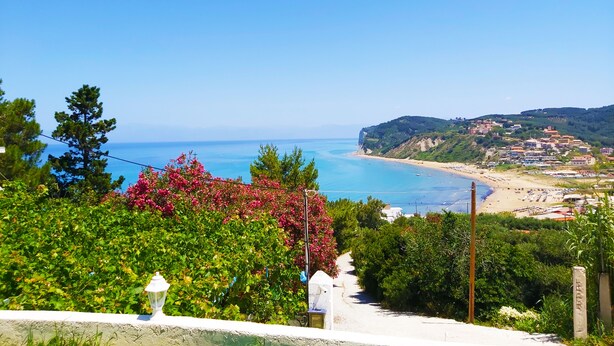 A coastal view with a beach, a path leading to the beach, and a hillside with buildings. - Gikas Apartments - Serenity Suite (Corfu)