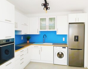 Une cuisine moderne avec des meubles blancs, un pare-feu bleu, un réfrigérateur en acier inoxydable, un lave-vaisselle encastré et un lave-linge.