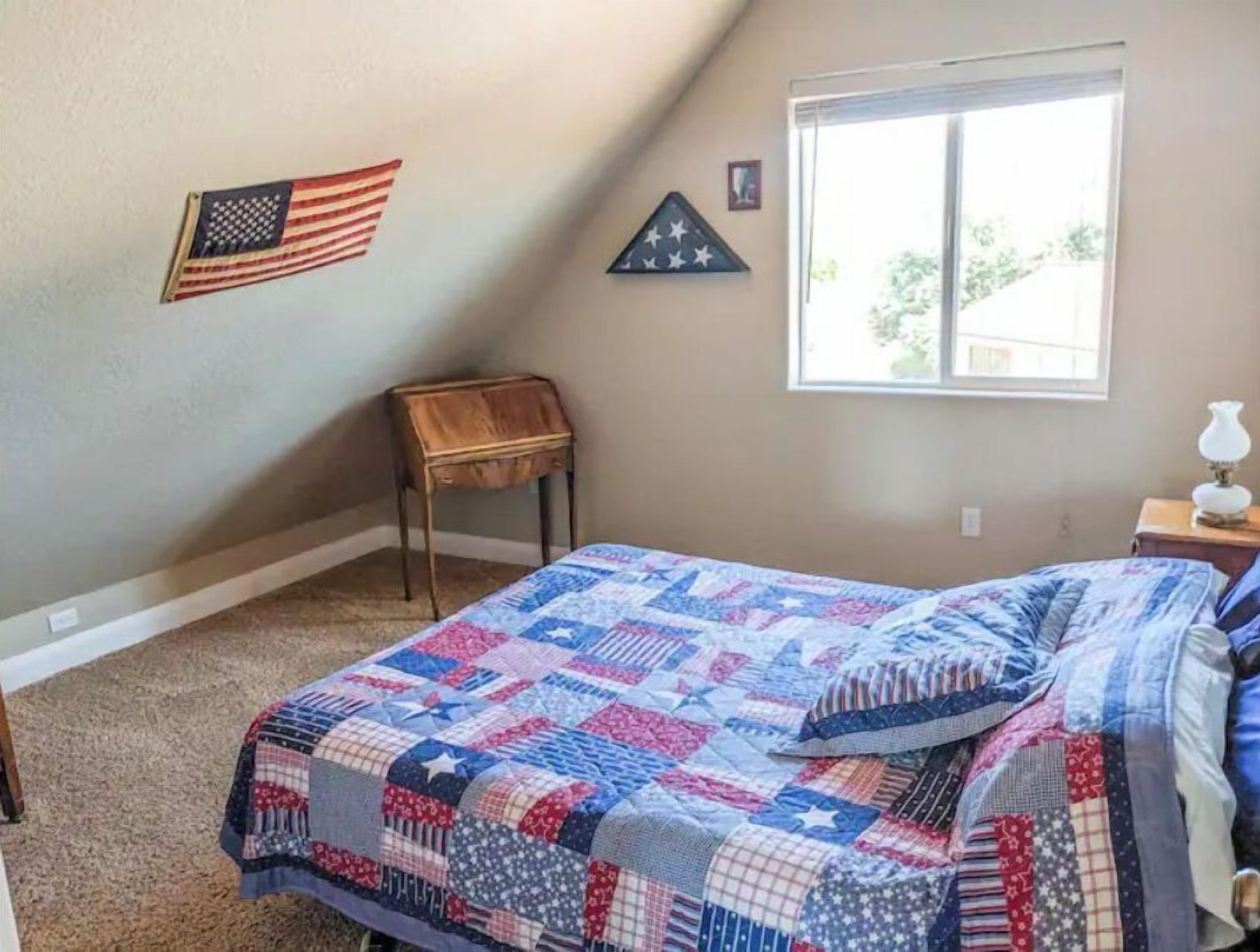 A bedroom with a bed covered in a patchwork quilt, a small wooden chair, a window with a view, and an American flag on the wall.