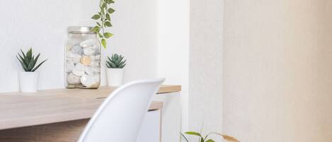 Une pièce minimaliste avec un bureau en bois, une chaise blanche et un mur blanc. On y trouve deux petites plantes en pot sur le bureau et une plante suspendue au-dessus.
