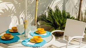 A round table with blue placemats and yellow bowls, surrounded by white wicker chairs, set outdoors with a palm tree and plants in the background. - Casa Indaiá - Caraguatatuba (Caraguatatuba)