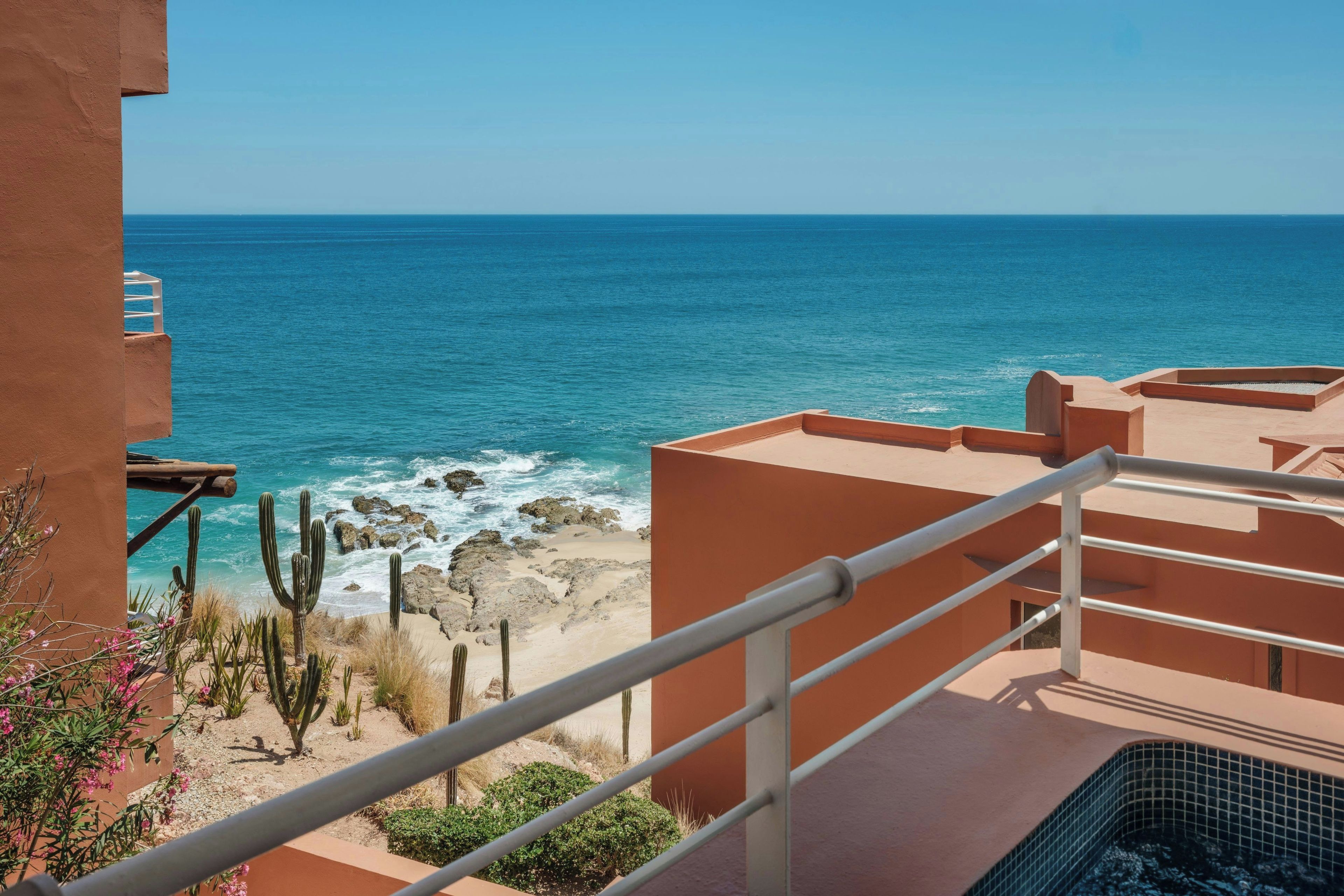 Une vue sur la côte depuis un balcon doté d’une balustrade, donnant sur la mer et une plage de sable où poussent des cactus.