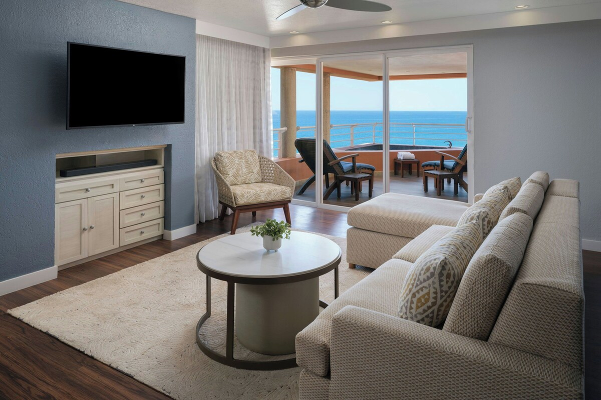 A modern living room with a flat-screen TV, a sofa, a coffee table, and a view of the ocean through a sliding glass door.