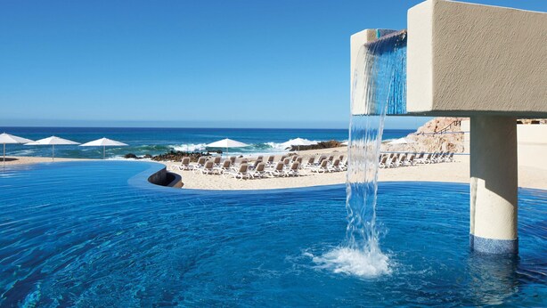 Una piscina infinita con cascada, sillas de playa y sombrillas.