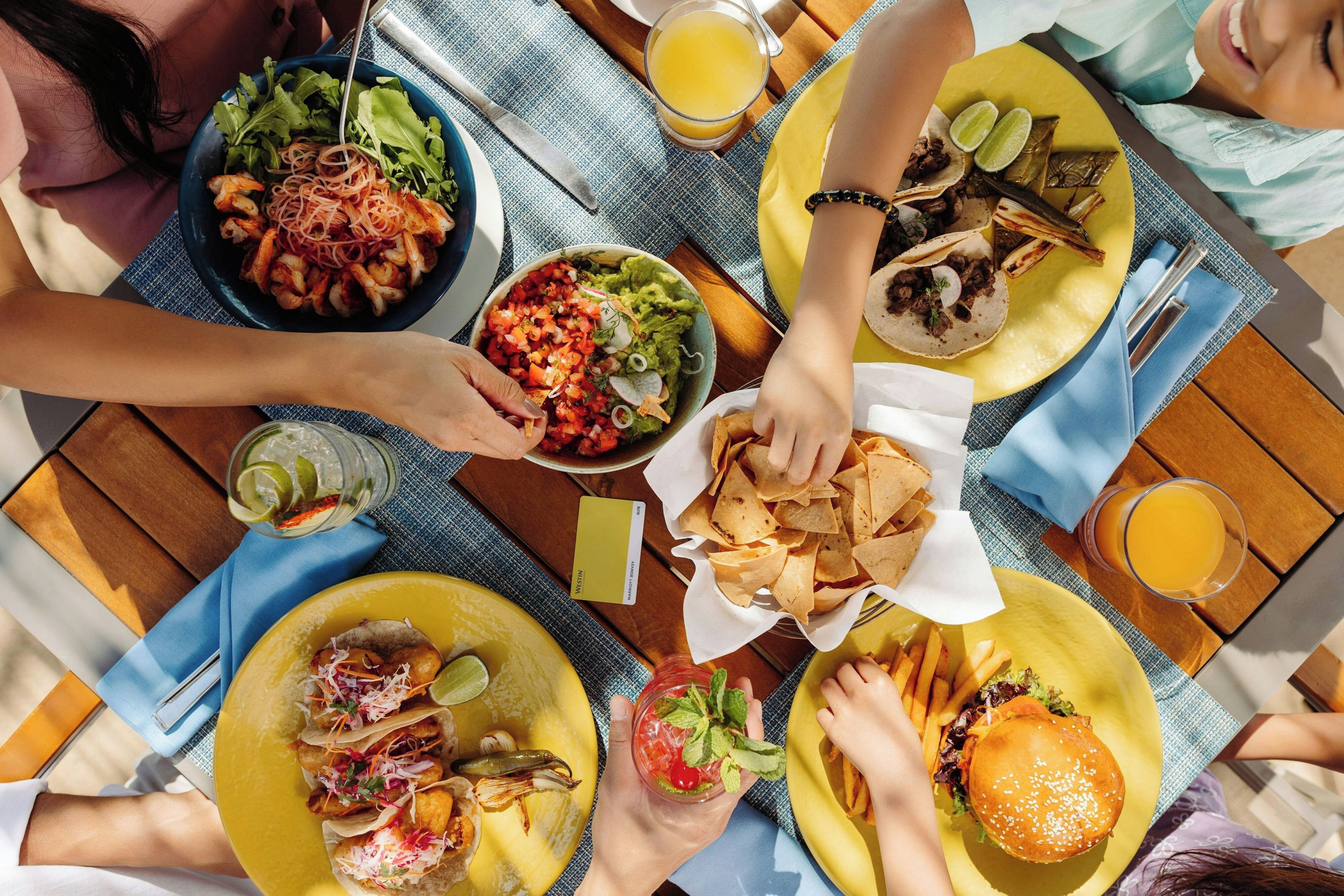 Eine Gruppe von Menschen speist draußen und genießt dabei verschiedene Gerichte, darunter Tacos, einen Burger und einen Salat.