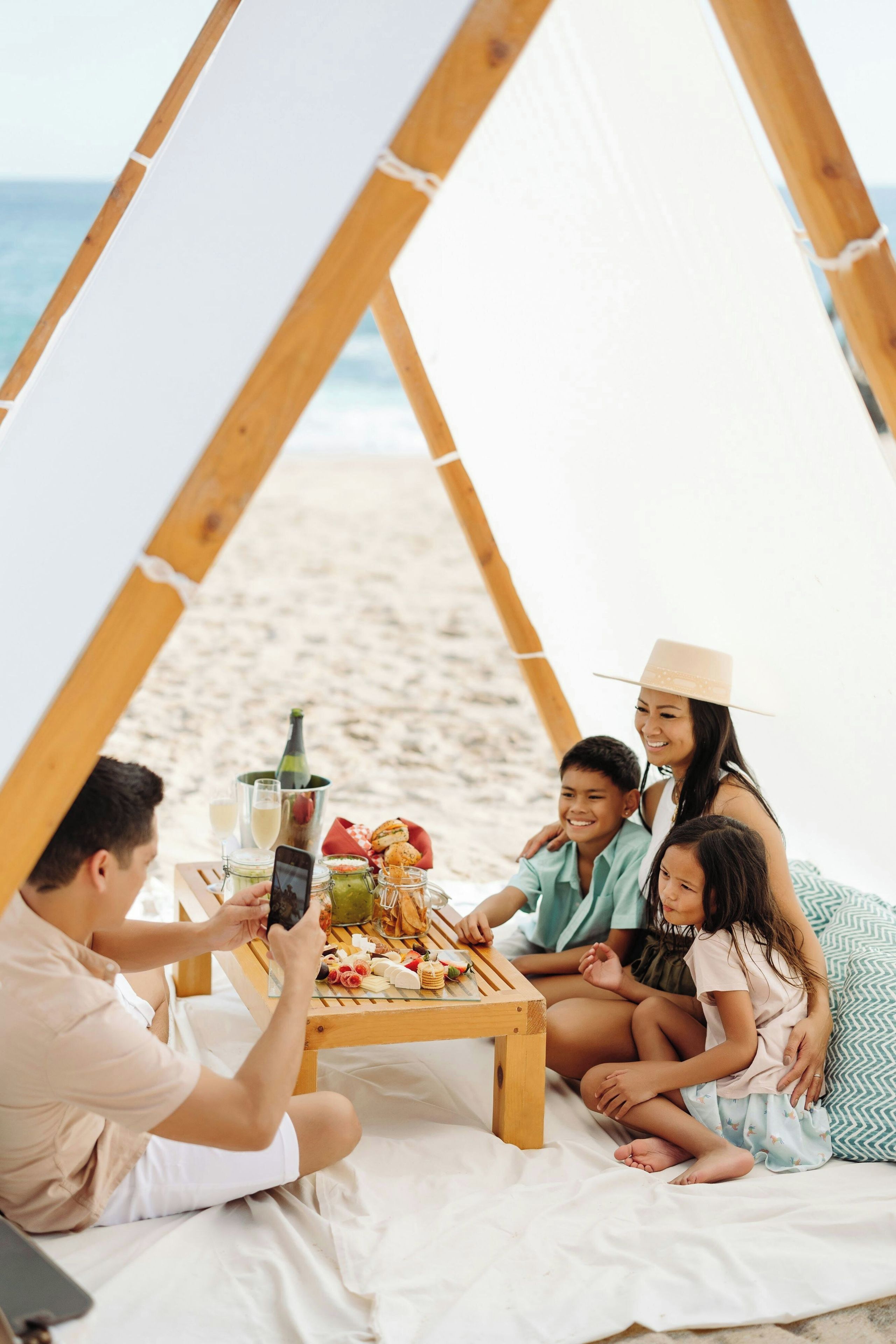 Una familia disfrutando de un picnic en la playa, con una estructura de madera y una mesa lista con comida y bebidas.