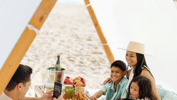 Una familia disfrutando de un picnic en la playa, con una estructura de madera y una mesa lista con comida y bebidas.