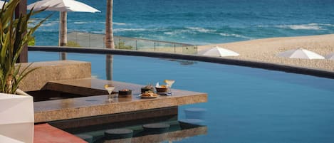 An infinity pool with a view of the ocean, palm trees, and beach umbrellas.