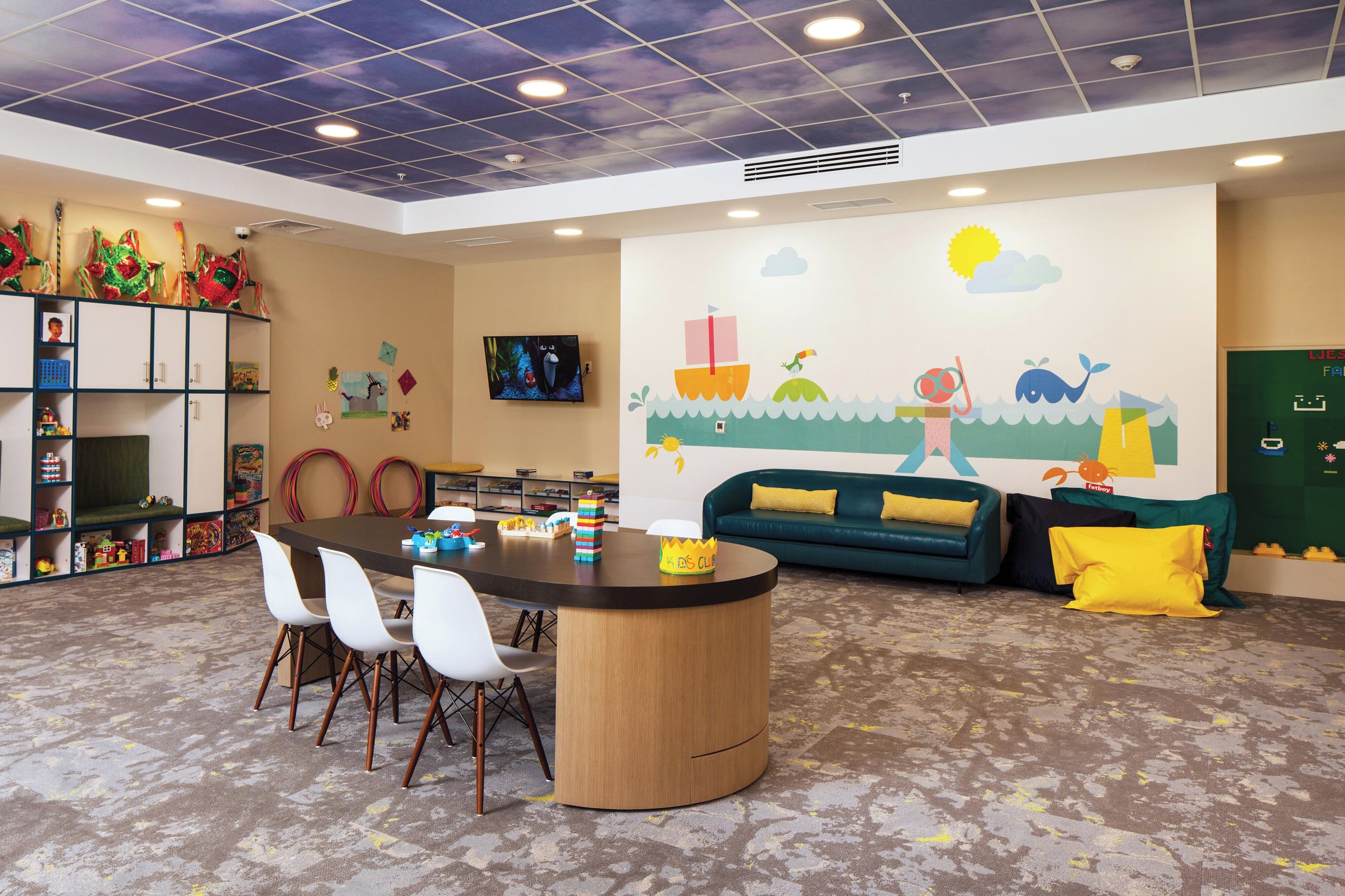 A children's playroom with a round table, chairs, a sofa, and a wall mural.