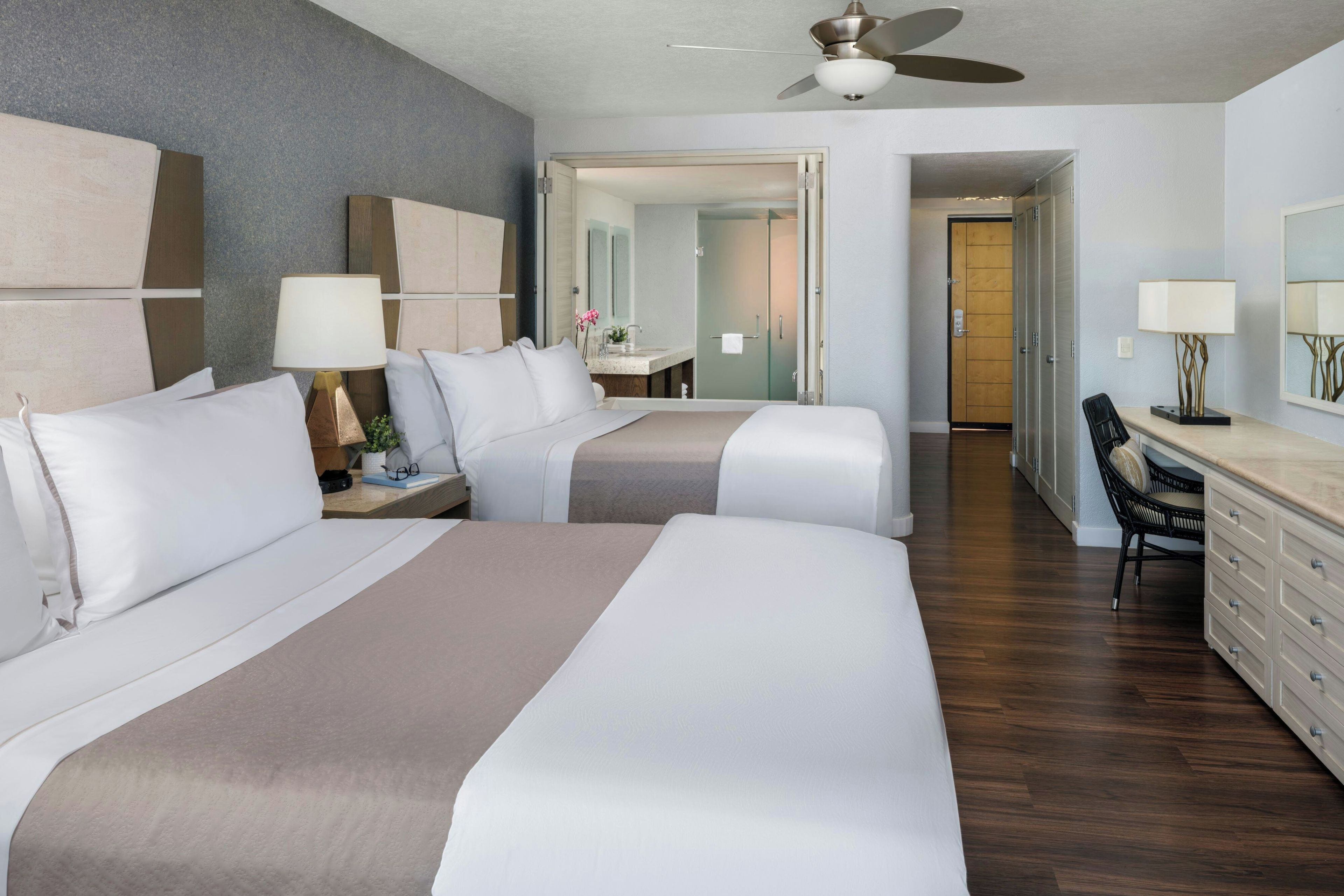 A hotel room with two beds, a desk, a chair, a ceiling fan, and a bathroom visible through an open door.