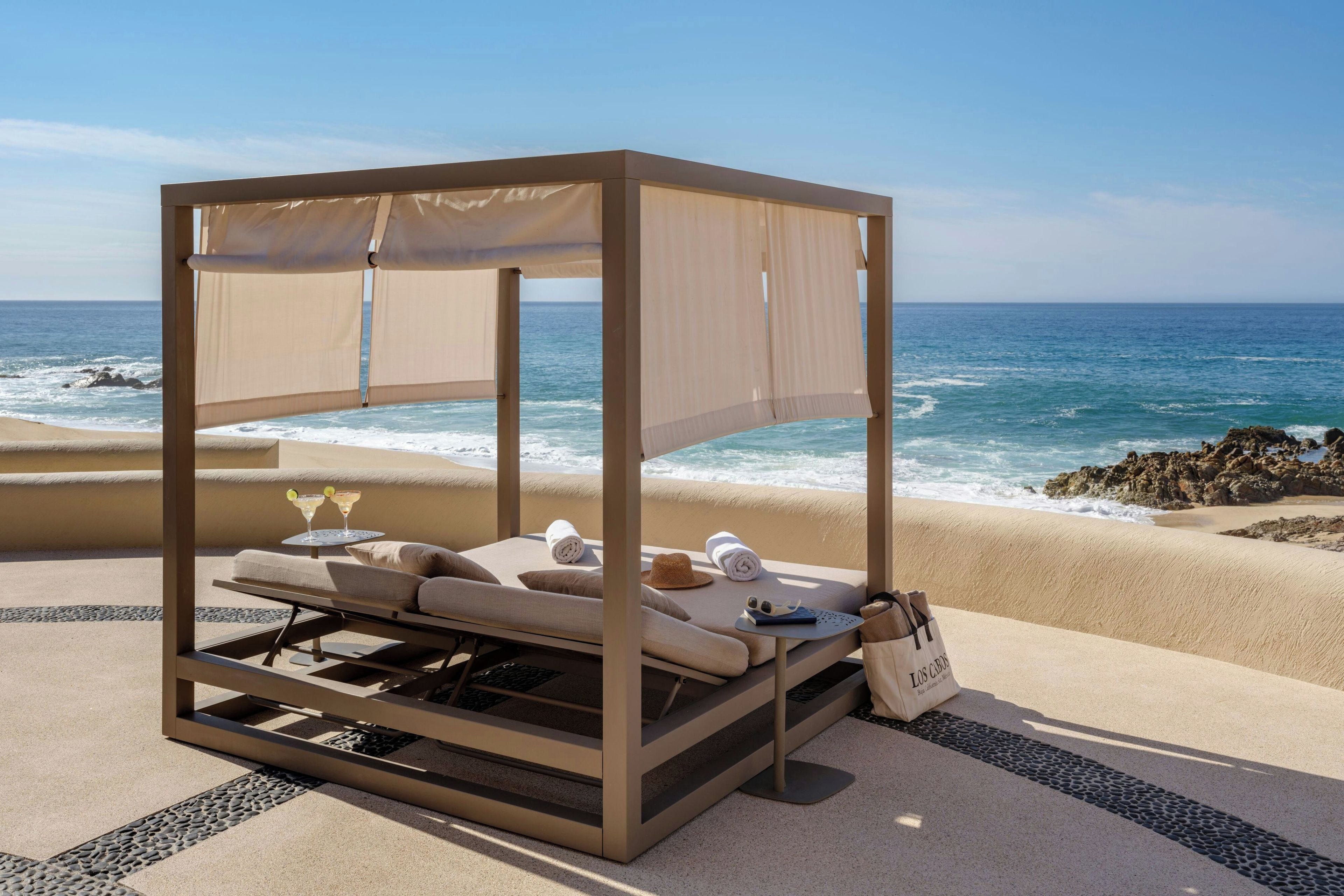 A beachside lounge with a canopy, lounge chairs, and a table with drinks.
