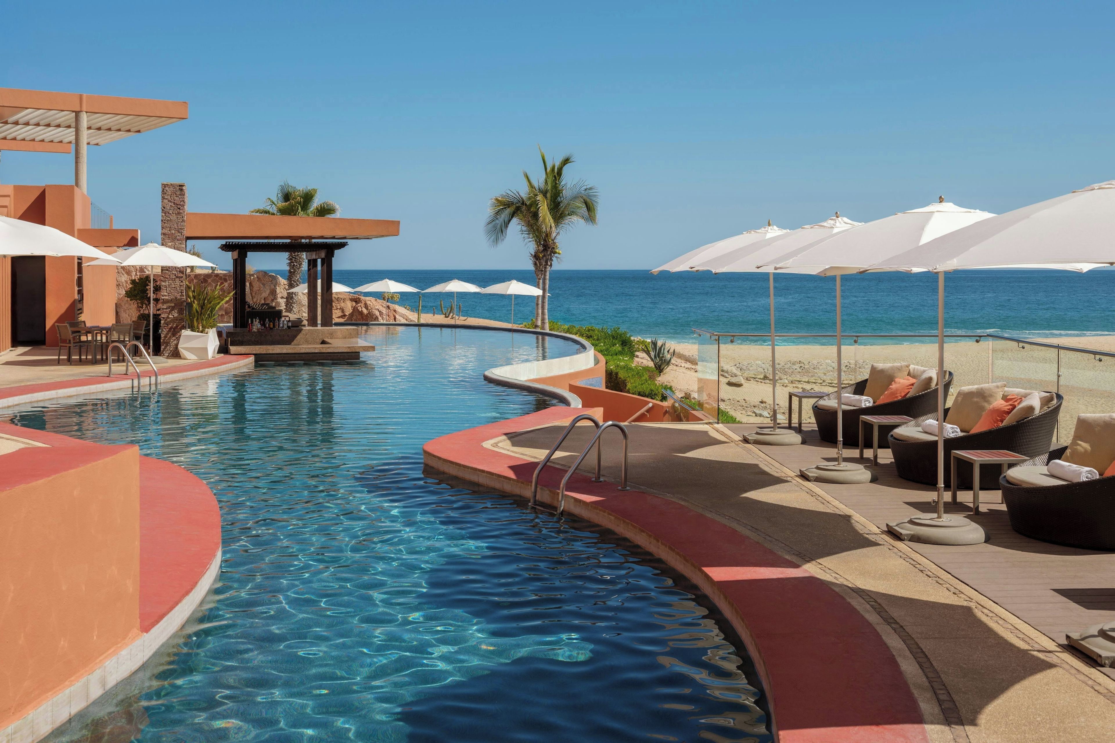 A pool area with a view of the sea, lounge chairs, and umbrellas.