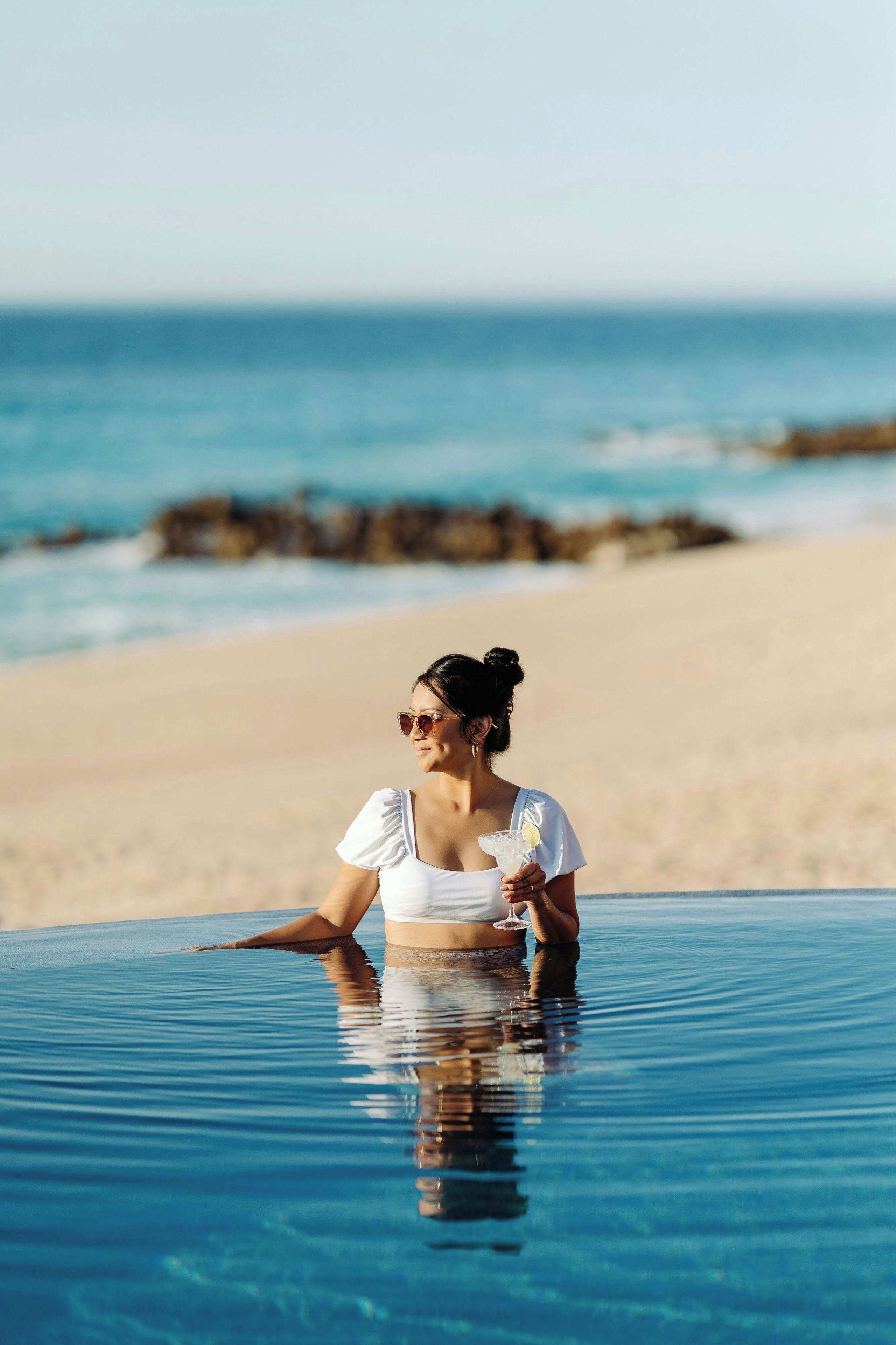 Una persona con remera blanca sentada en una pileta transparente con vista a la playa y al océano.