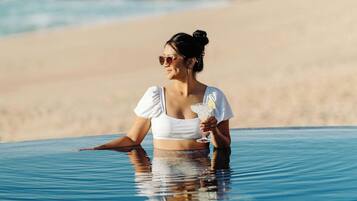 Una persona con remera blanca sentada en una pileta transparente con vista a la playa y al océano.