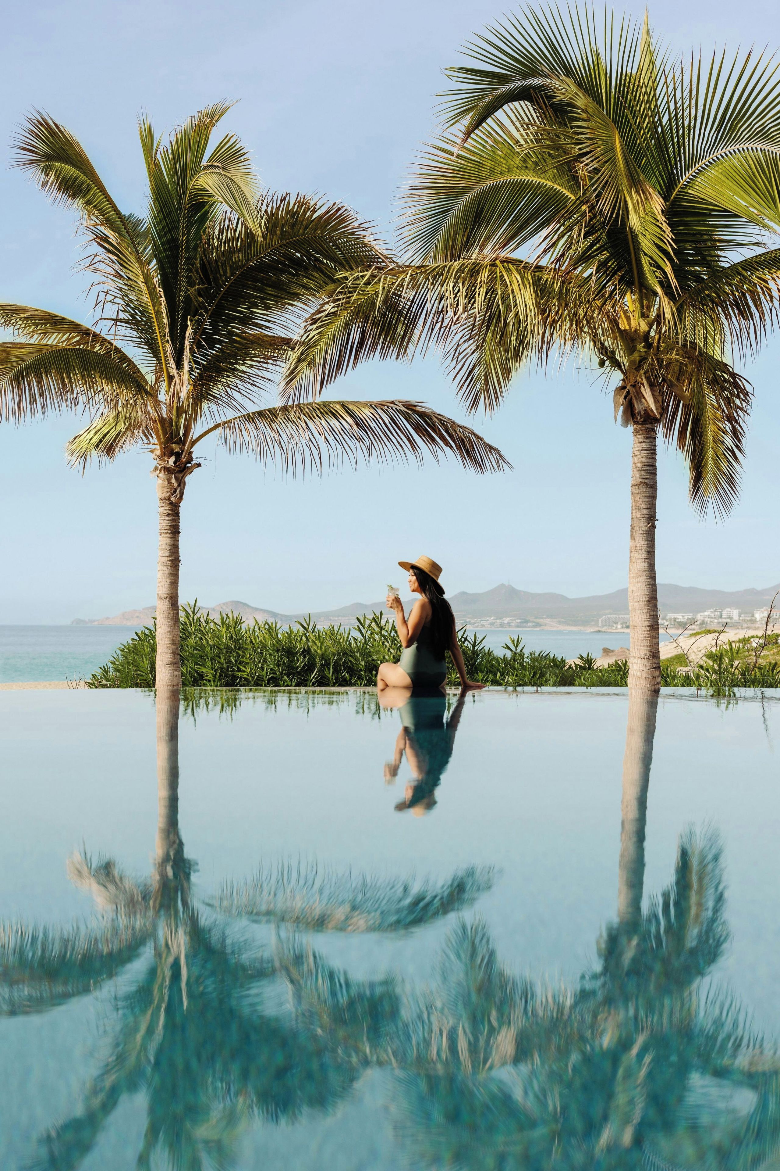 Una persona sentada junto a una piscina infinita, con palmeras y vistas al mar y las montañas.