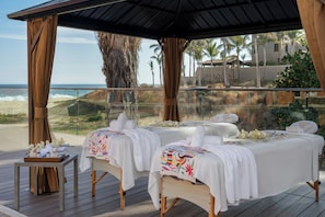 Un spa frente al mar con dos camillas de masaje, una terraza de madera y vista al océano.