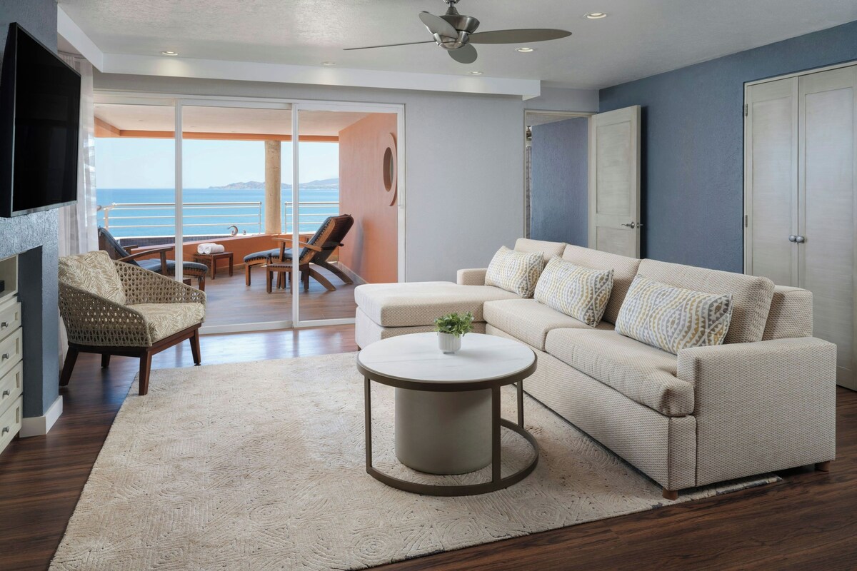 A modern living room with a sofa, a coffee table, and a TV. There is a balcony with a view of the sea.