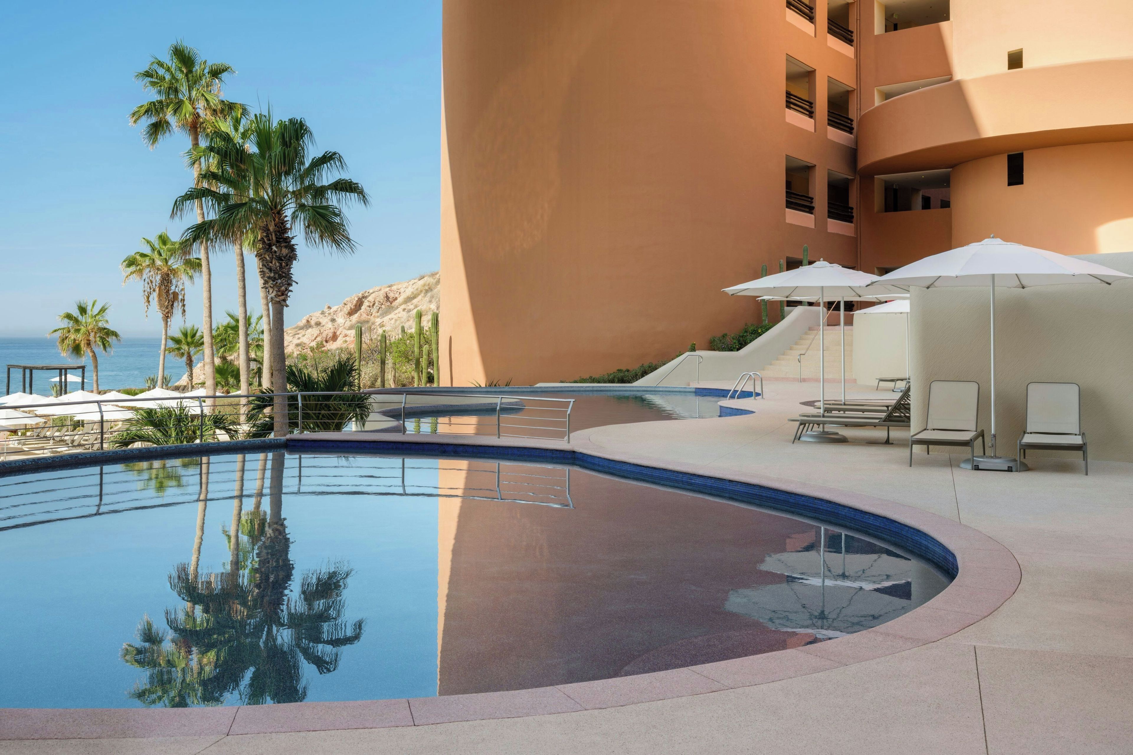 A swimming pool with lounge chairs and umbrellas, surrounded by palm trees and a building.