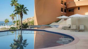 A swimming pool with lounge chairs and umbrellas, surrounded by palm trees and a building.