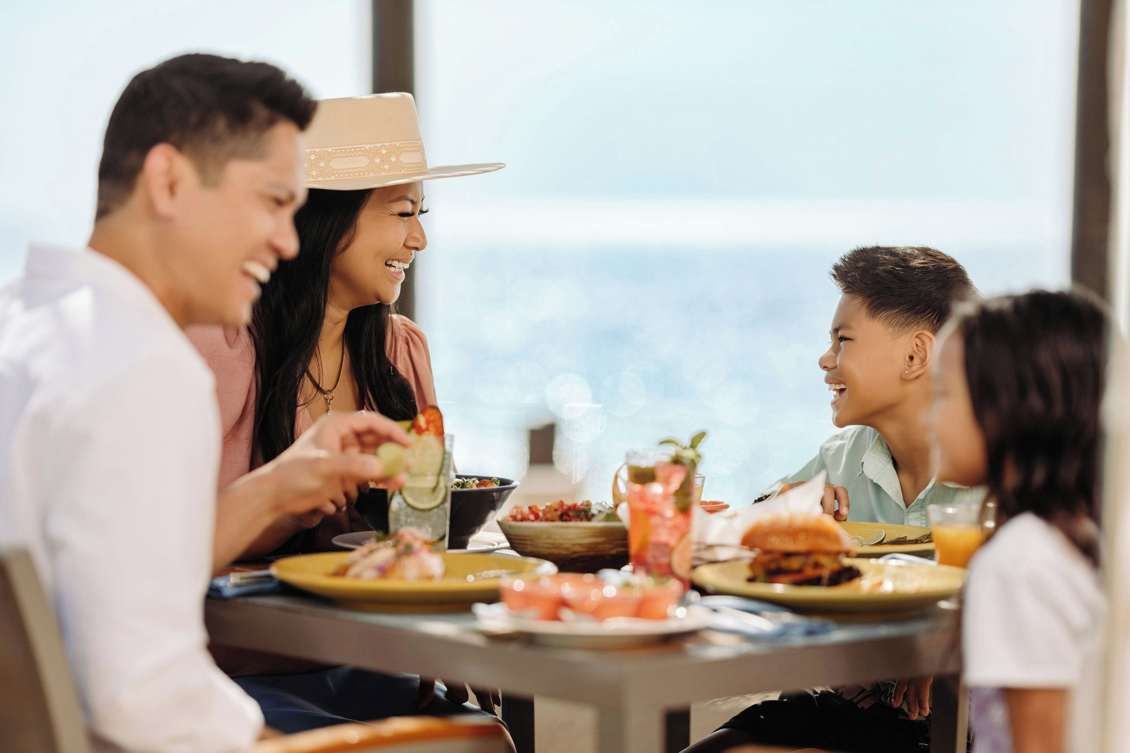 Una familia cenando en una mesa con diversos platos y bebidas, con vistas a un paisaje pintoresco.