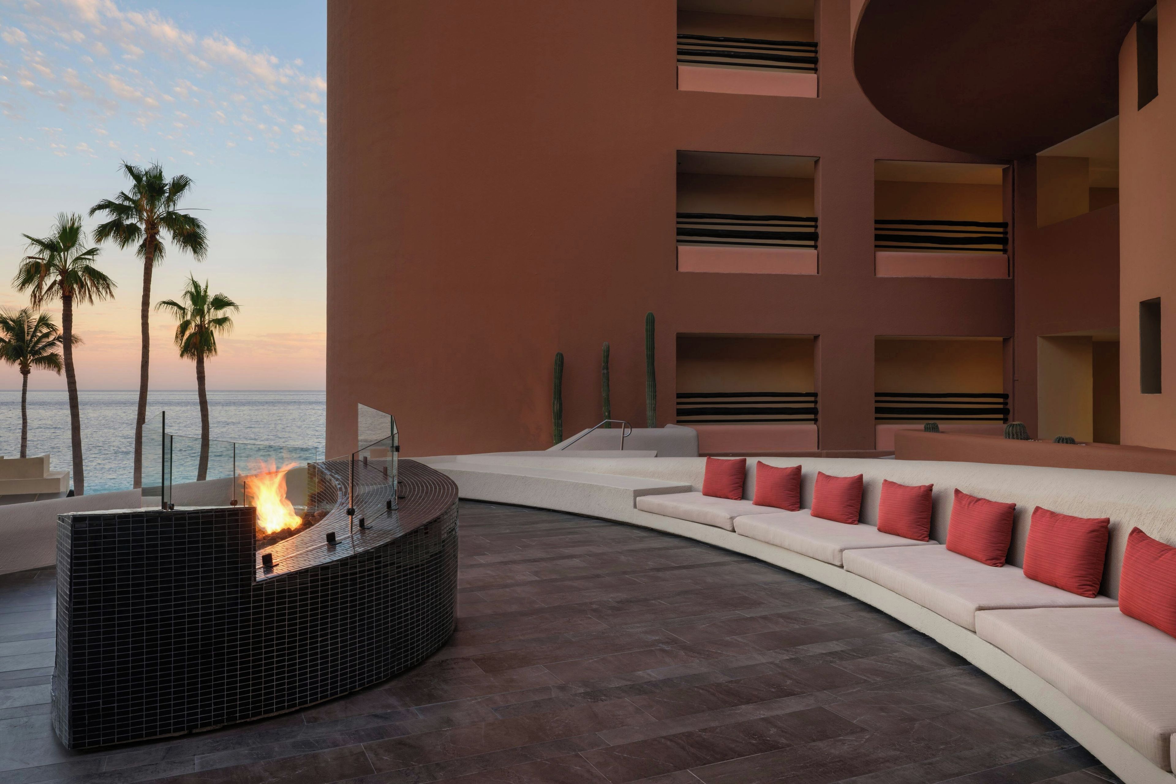 A curved outdoor seating area with a fire feature, palm trees, and a view of the ocean.