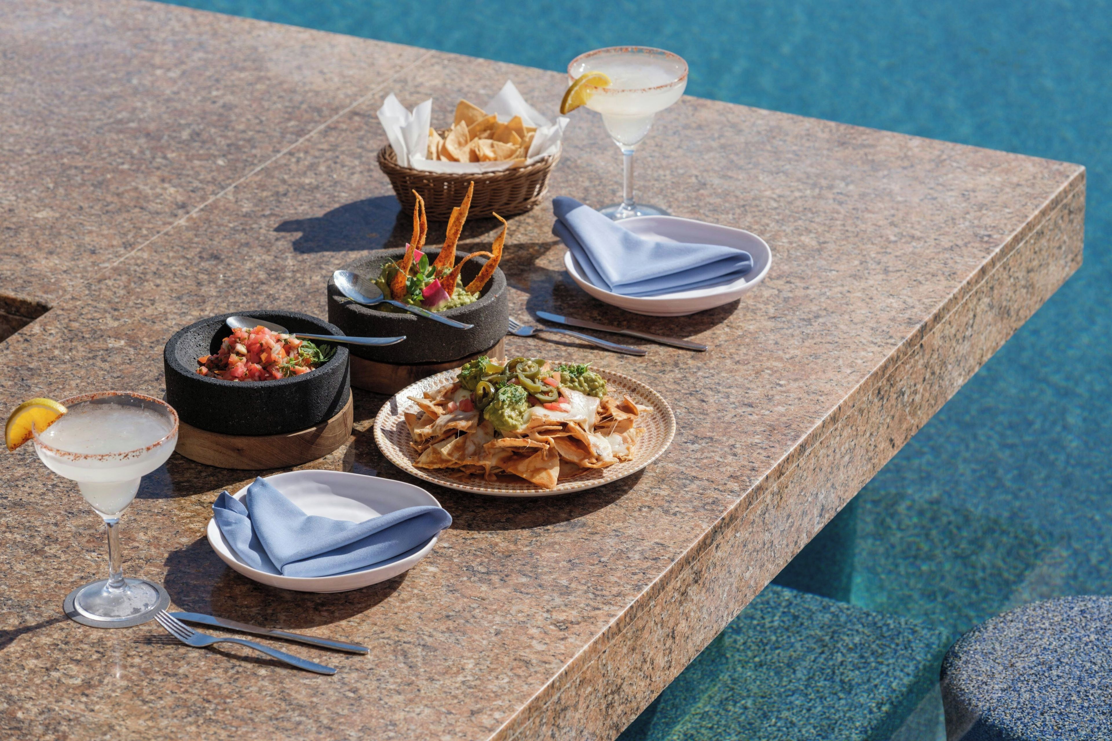 A dining table with a variety of dishes, including nachos, a margarita, and a bowl of salsa.