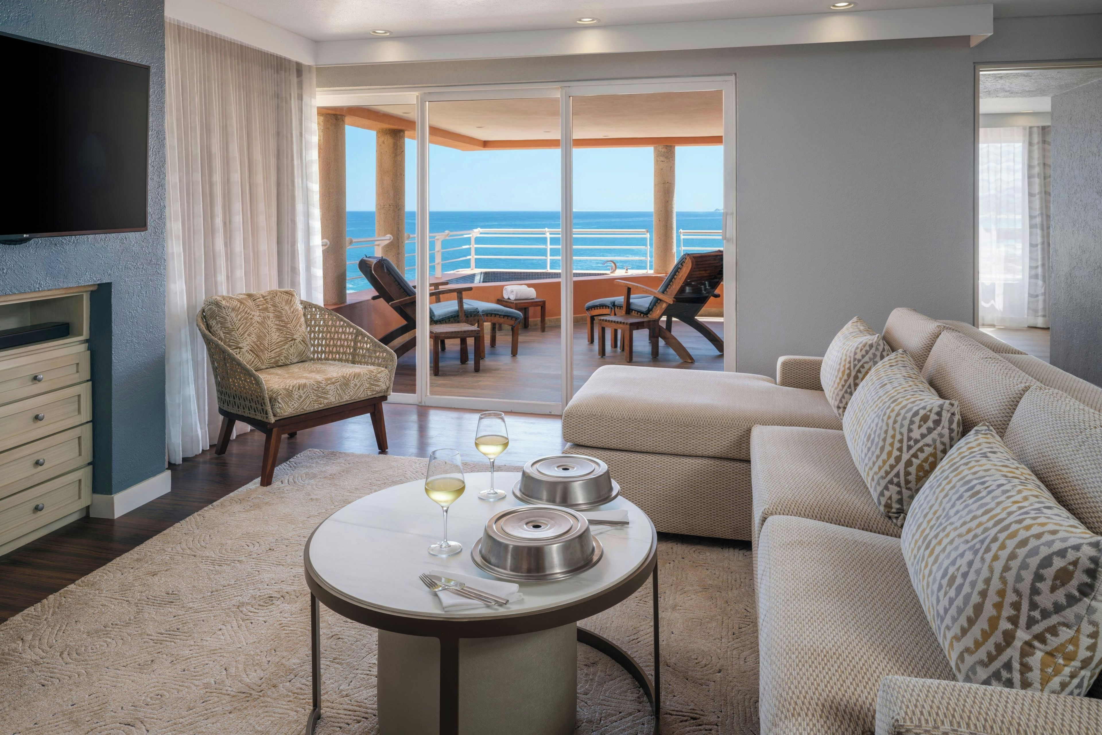 A modern living room with a sofa, a dining table with chairs, a flat-screen TV, and a view of the ocean through a sliding glass door.