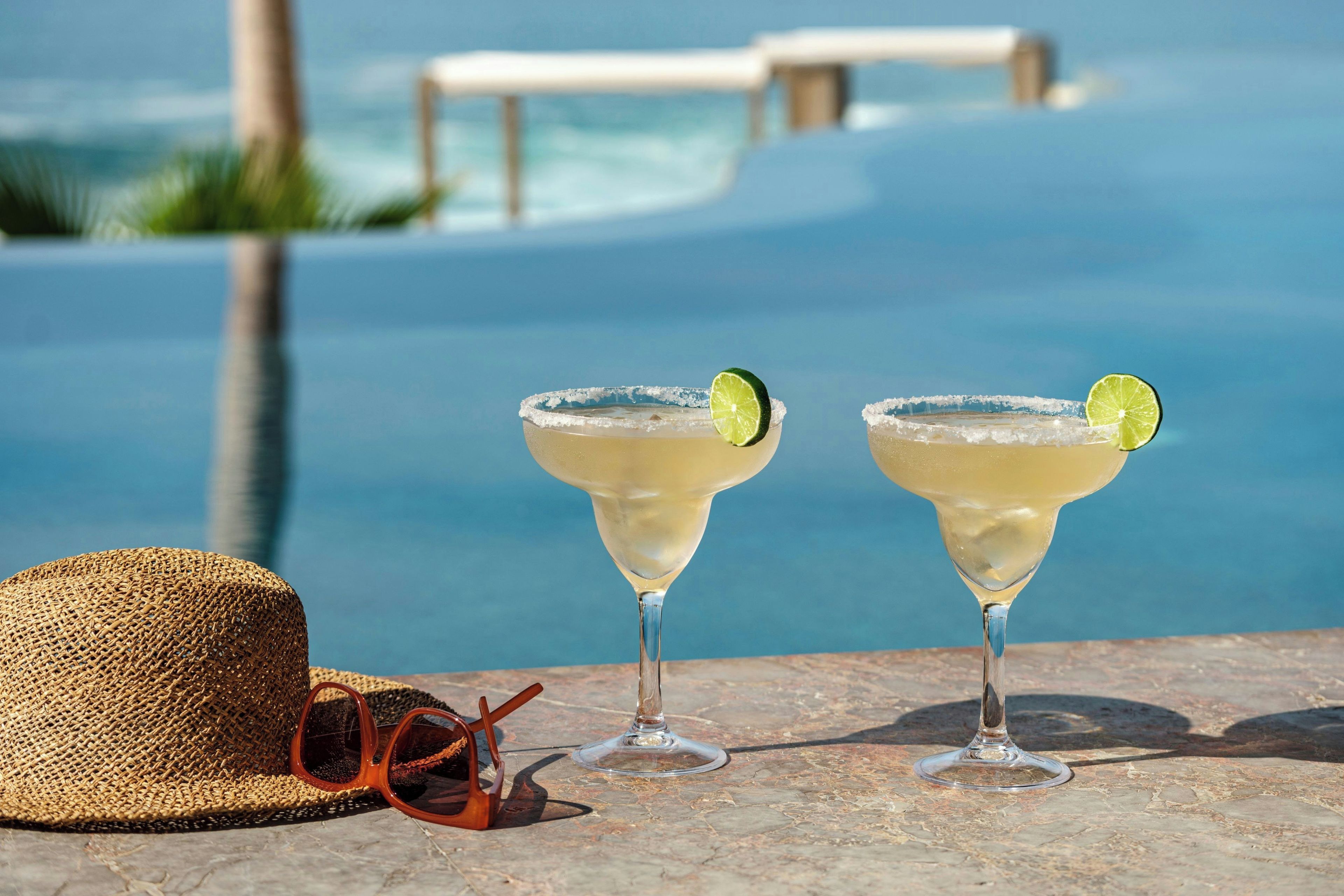 Two margaritas with lime garnish on a stone table by a poolside with a straw hat and sunglasses nearby.