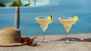 Two margaritas with lime garnish on a stone table by a poolside with a straw hat and sunglasses nearby.