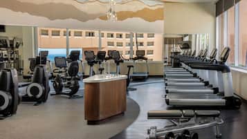 Ett gym med konditionsmaskiner, styrketräningsutrustning och ett stort fönster med utsikt över byggnader.