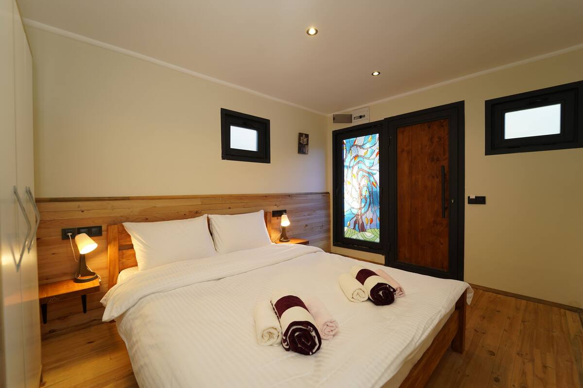 Une chambre d’hôtel avec une tête de lit en bois, un lit recouvert de draps blancs, deux lampes de chevet et une fenêtre donnant sur un vitrail.
