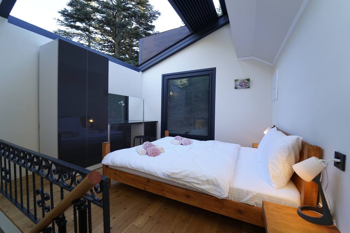 Une chambre avec un lit en bois, du linge de lit blanc, une table de chevet, une lampe, une fenêtre et une vue sur les arbres à l’extérieur.