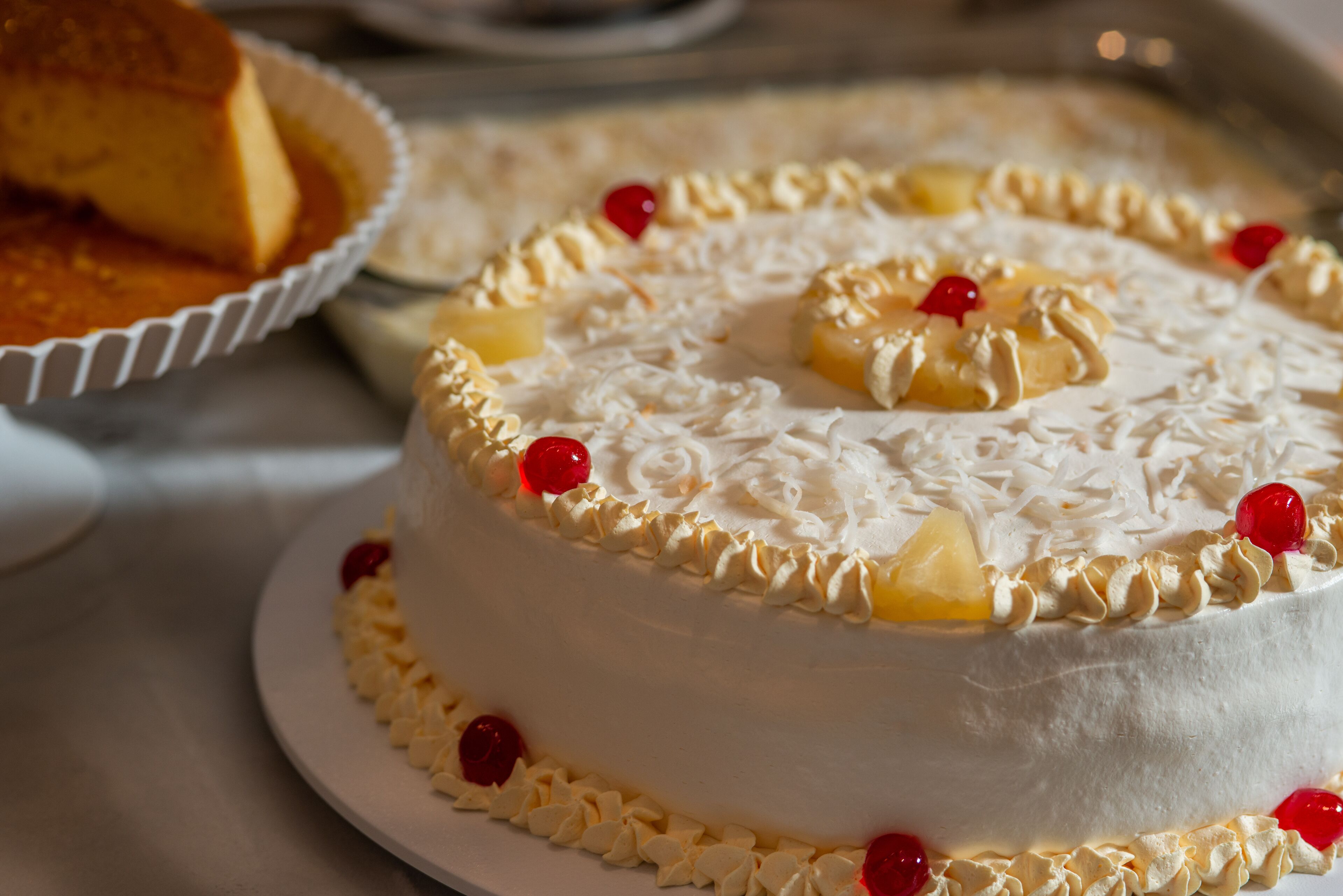 Um bolo com cobertura branca, pedaços de abacaxi e cerejas vermelhas sobre um prato branco.