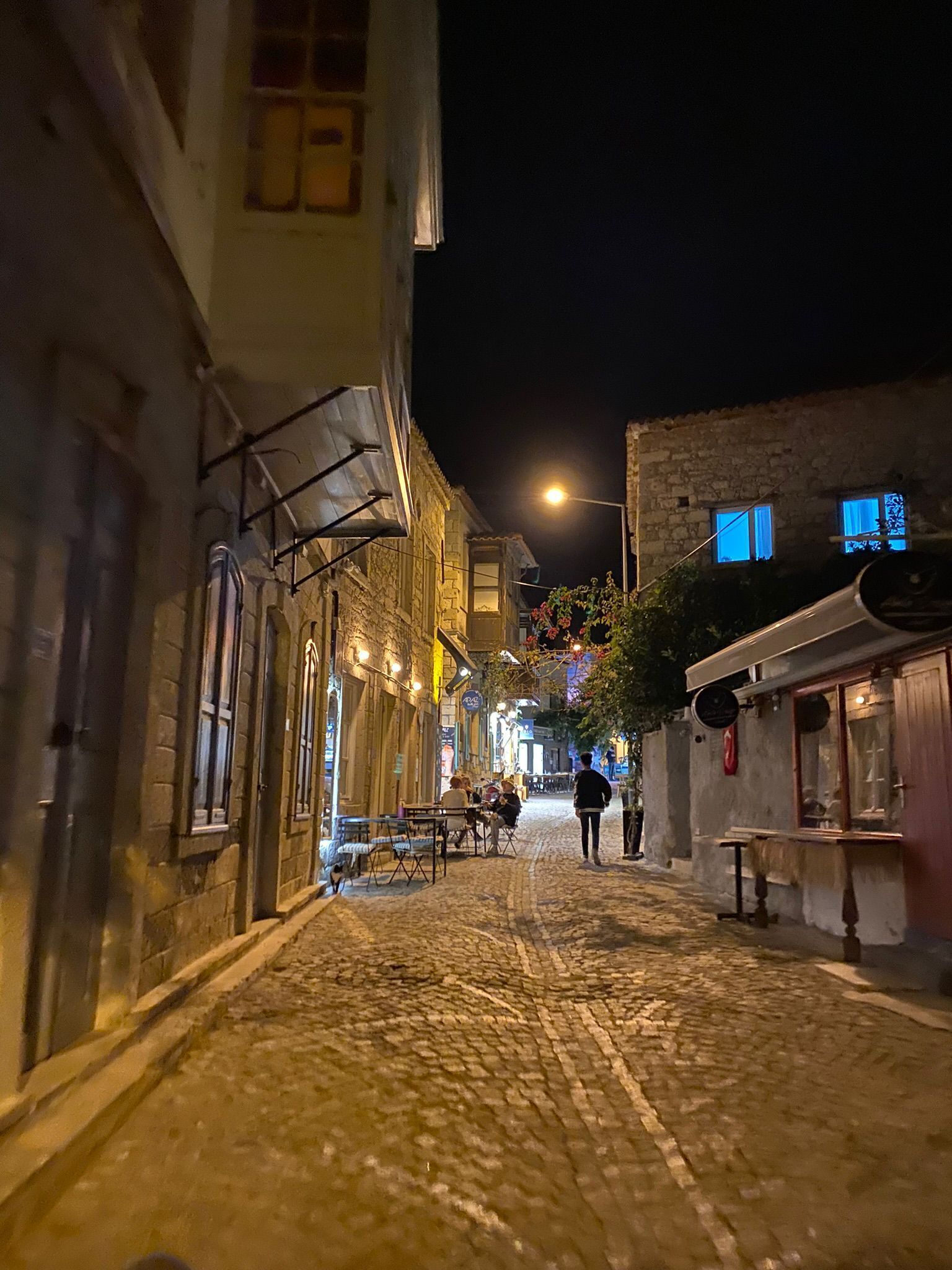 Eine Kopfsteinpflasterstraße bei Nacht mit Gebäuden an beiden Seiten, einige mit Außenbestuhlung, wenige Personen sind sichtbar.