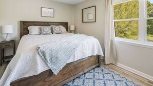Une chambre à coucher avec un lit en bois, deux tables de chevet avec des lampes, un tapis à motifs et une fenêtre donnant sur un paysage verdoyant.