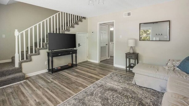 Un salon avec un escalier, un téléviseur à écran plat posé sur un meuble, un canapé, un tapis à motifs et une petite table avec une lampe.