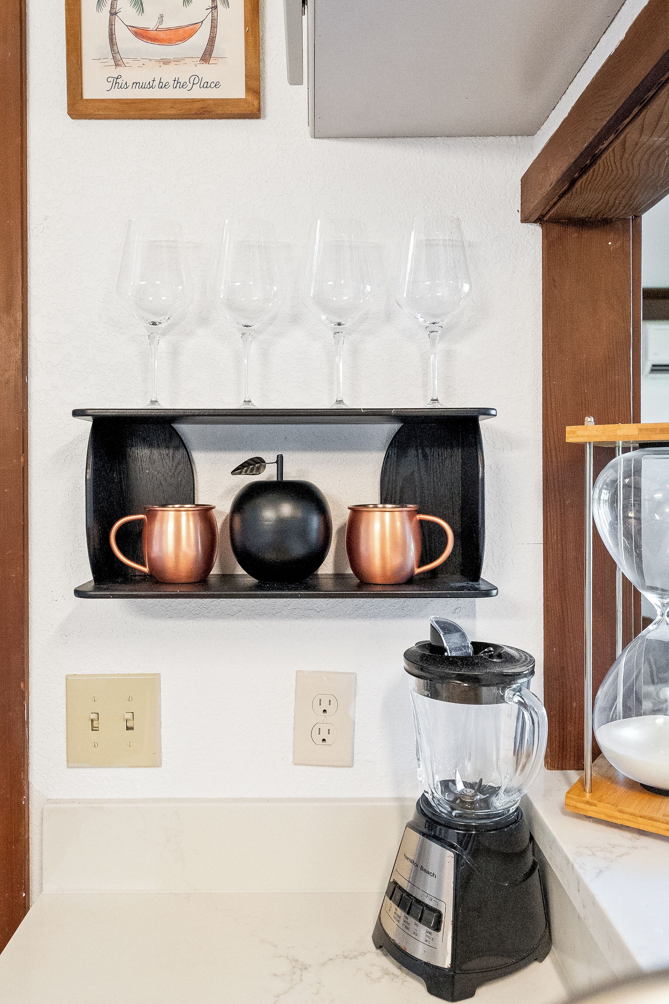 A kitchen with a black countertop, a blender, a shelf with two copper mugs, and a framed picture above.