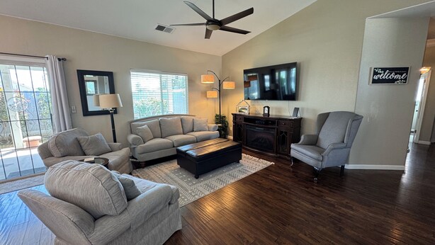 A spacious living room with a large sofa, a coffee table, and a fireplace. There is a ceiling fan, a TV mounted on the wall, and a 'welcome' sign.